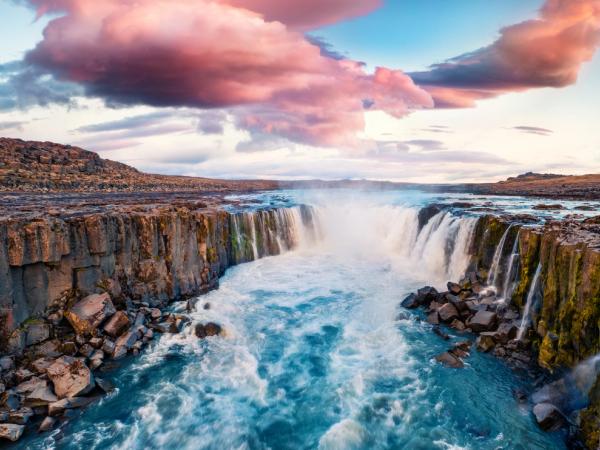 Una poderosa cascada se precipita en un río azul y turbulento, rodeado de oscuros acantilados de basalto, bajo un cielo con nubes rosas y moradas.