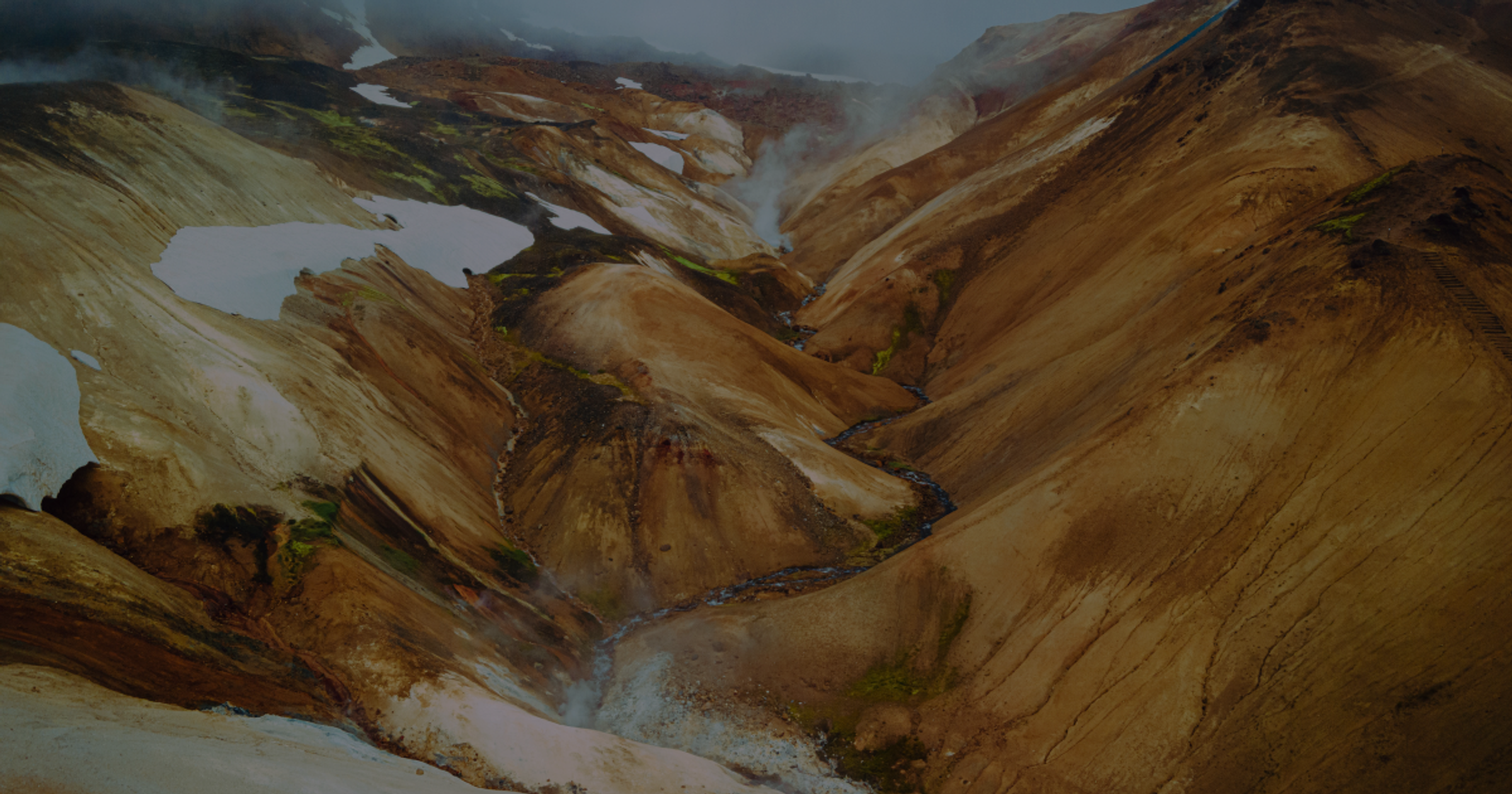 A volcanic valley with multicolored slopes, patches of snow, a winding river, and steam rising.