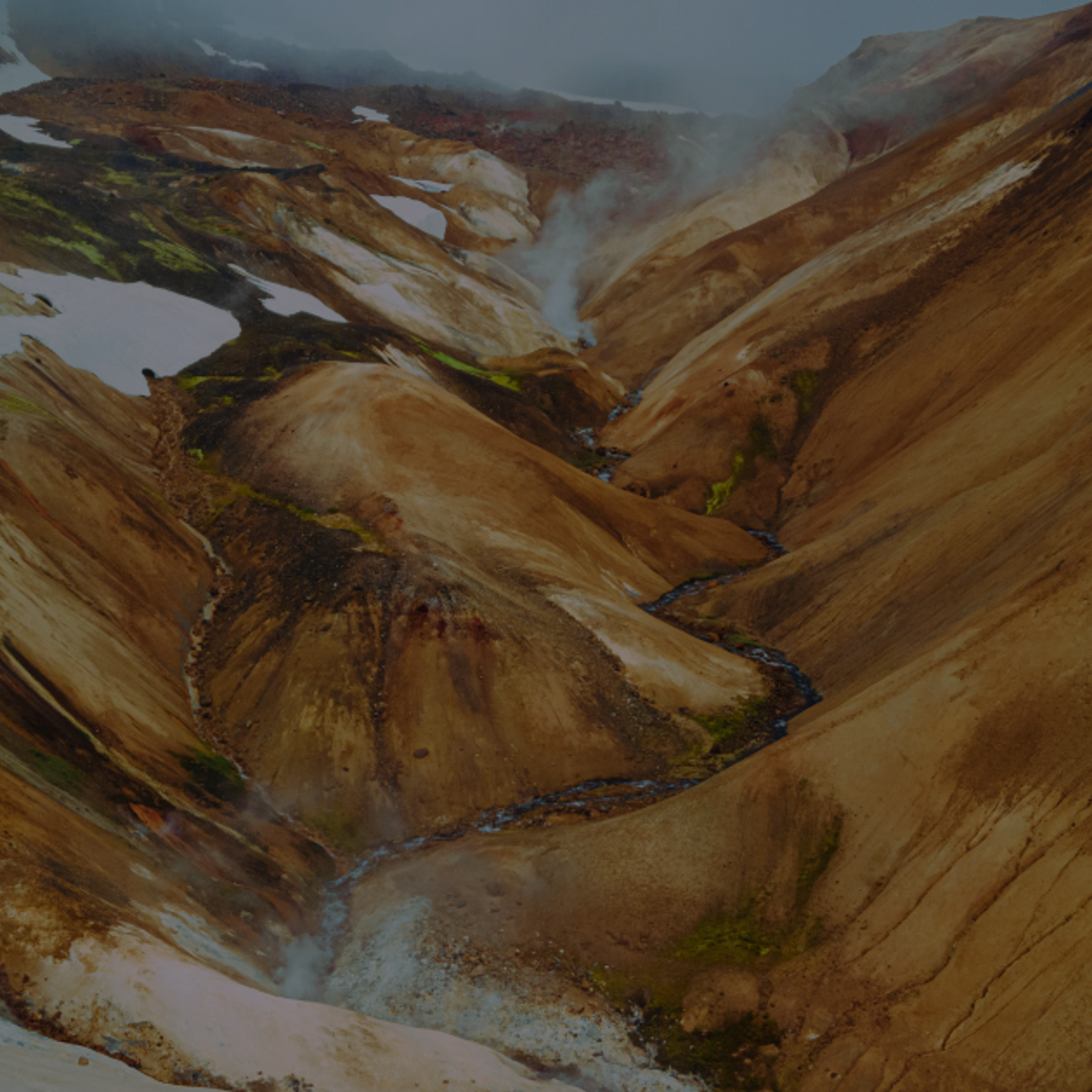 A volcanic valley with multicolored slopes, patches of snow, a winding river, and steam rising.