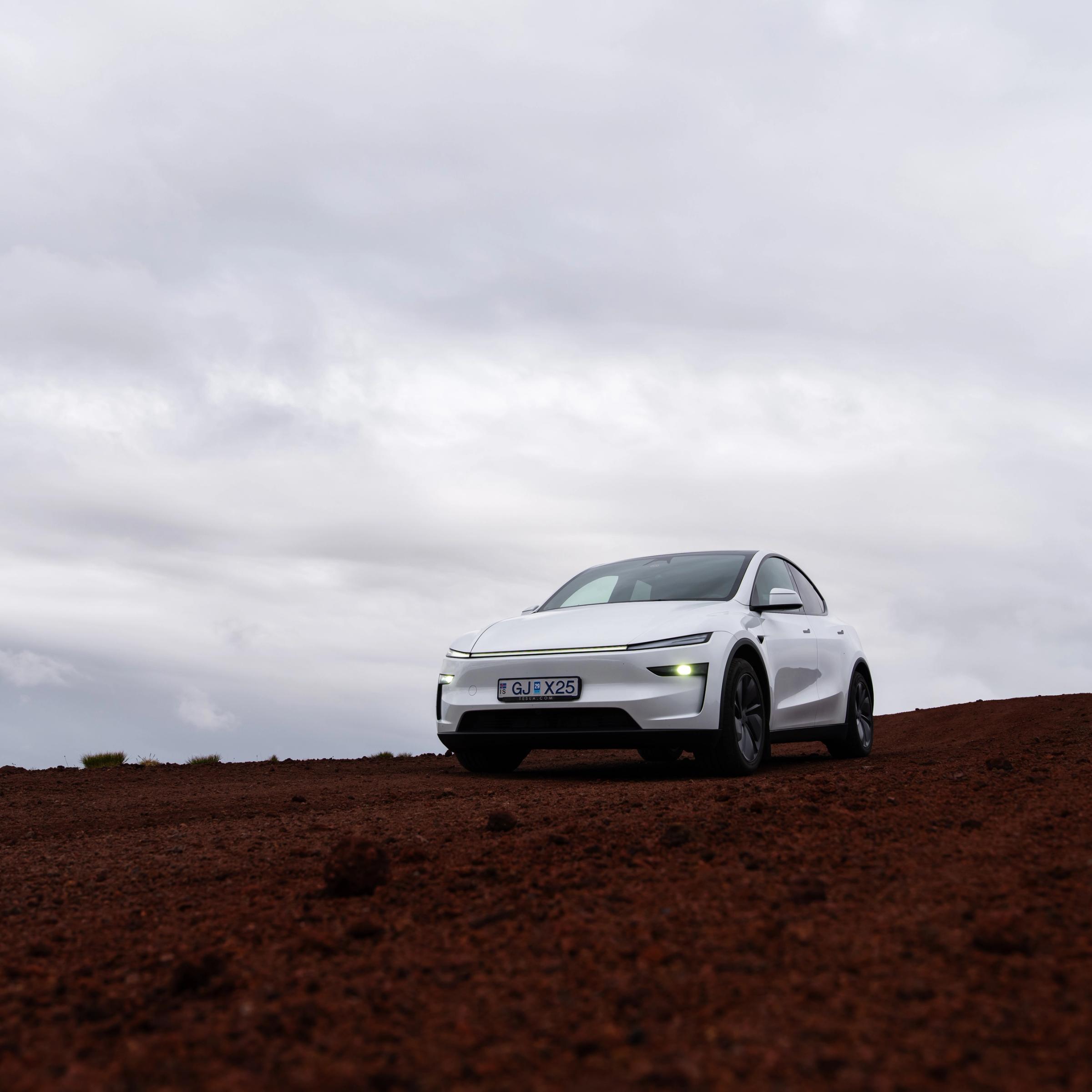 A white electric car is parked on a reddish-brown dirt road under a cloudy sky.