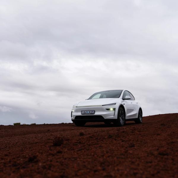 A white electric car is parked on a reddish-brown dirt road under a cloudy sky.