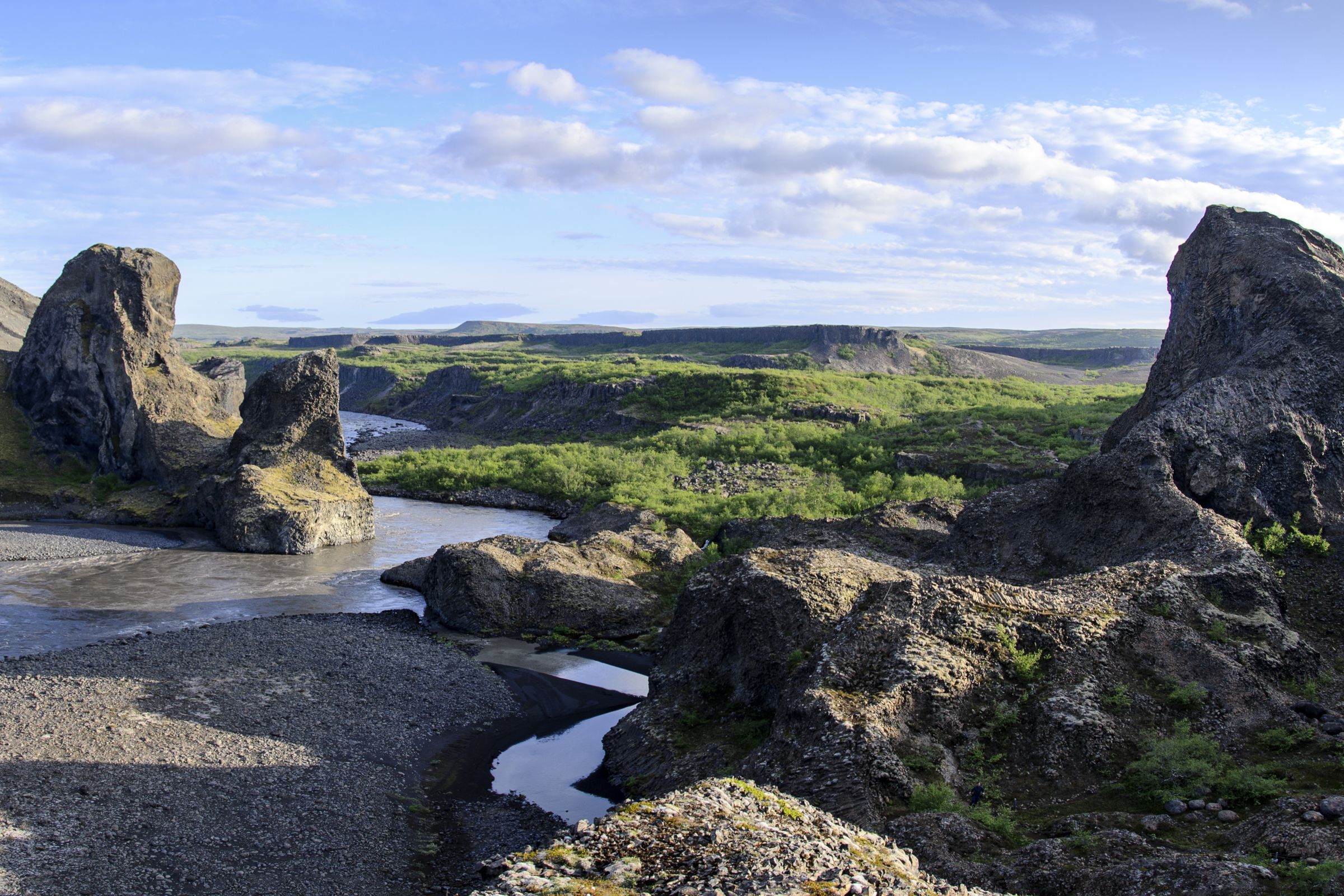 Rocky valley with a river, prominent rock formations, and green vegetation under a partly cloudy sky.