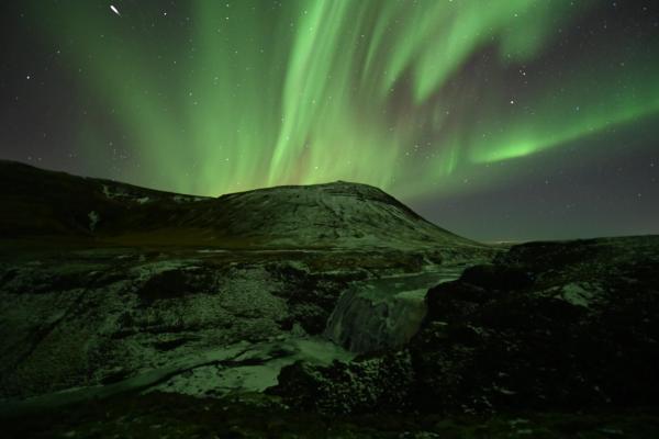 Vibrant green aurora borealis over a snowy mountain landscape with a waterfall.