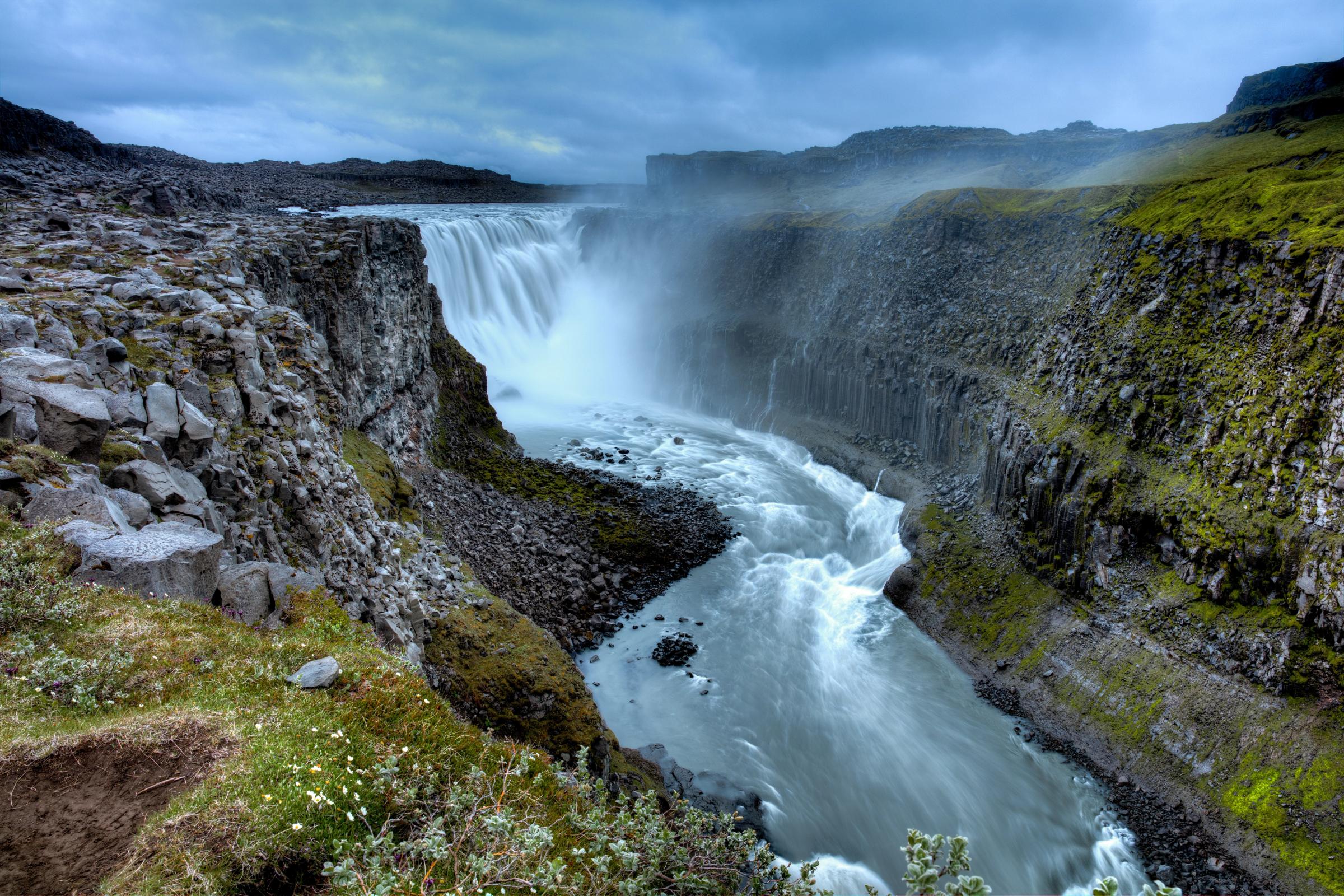 A powerful waterfall cascades into a deep, rocky canyon with green-tinged cliffs under a cloudy sky.