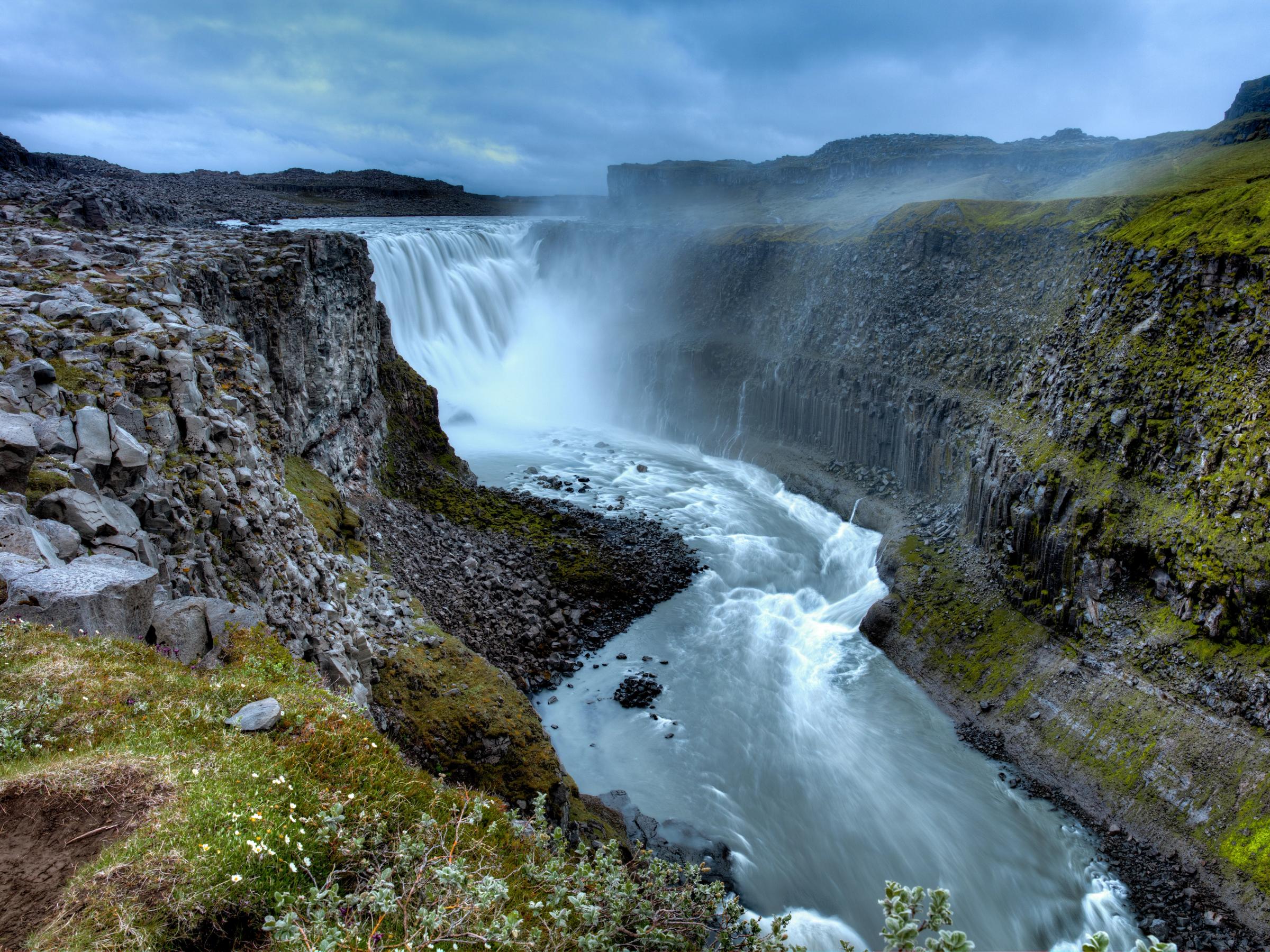 A powerful waterfall cascades into a deep, rocky canyon with green-tinged cliffs under a cloudy sky.