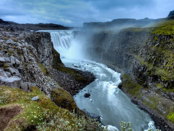 Una poderosa cascada cae en un profundo cañón rocoso, rodeado de acantilados de tono verdoso bajo un cielo nublado.