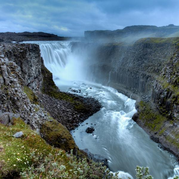 A wide waterfall plunges into a rocky canyon, with green moss on the cliffs under a cloudy sky.