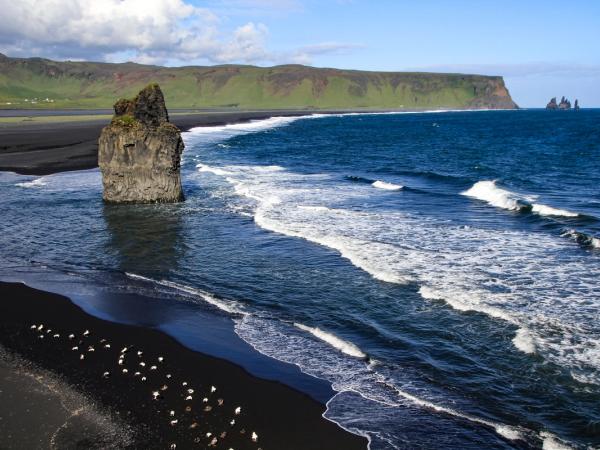 une plage de sable noir avec un gros rocher au milieu lors d'une journée ensoleillée