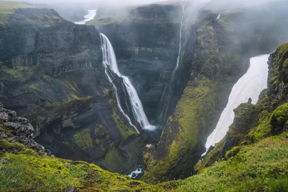 A deep canyon with multiple waterfalls, green mossy cliffs, dark rock, and a large snowfield under a foggy sky.