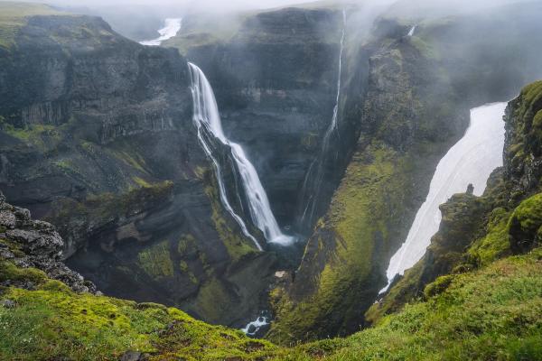 Misty canyon with two powerful waterfalls, dark rock cliffs, green moss, and a patch of snow.