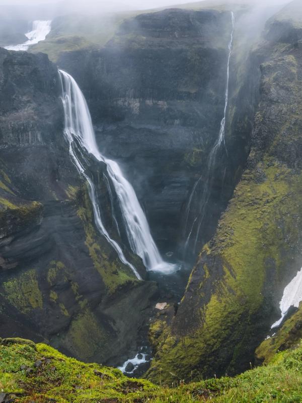 A deep canyon with multiple waterfalls, green mossy cliffs, dark rock, and a large snowfield under a foggy sky.