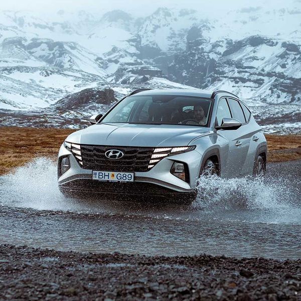 Silver Hyundai Tucson SUV splashing through a puddle with snowy mountains in the background.