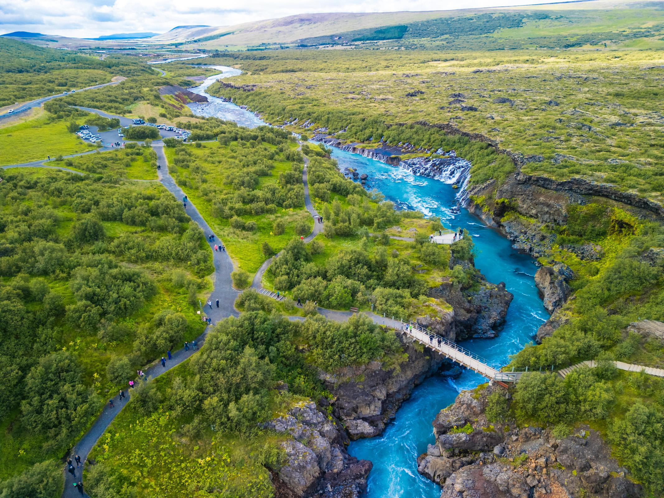Aerial view of a bright turquoise river with waterfalls and a pedestrian bridge, surrounded by green shrubland with walking paths and people.