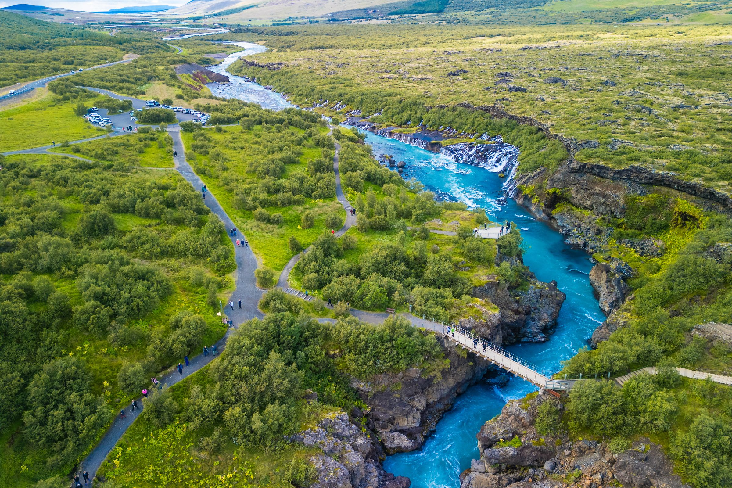 Aerial view of a bright turquoise river with waterfalls and a pedestrian bridge, surrounded by green shrubland with walking paths and people.