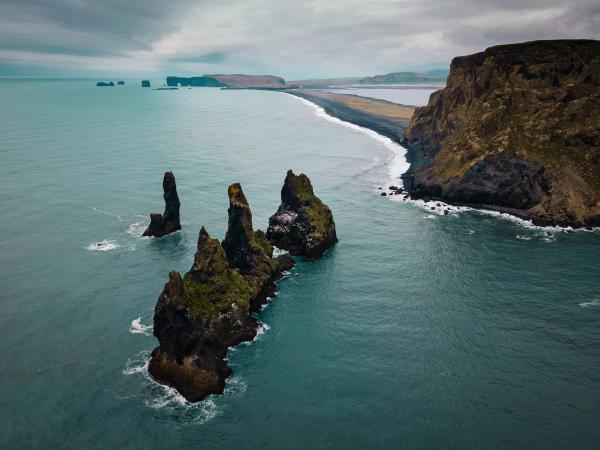Three jagged sea stacks in turquoise ocean water beside a black sand beach and dark cliffs under a cloudy sky.