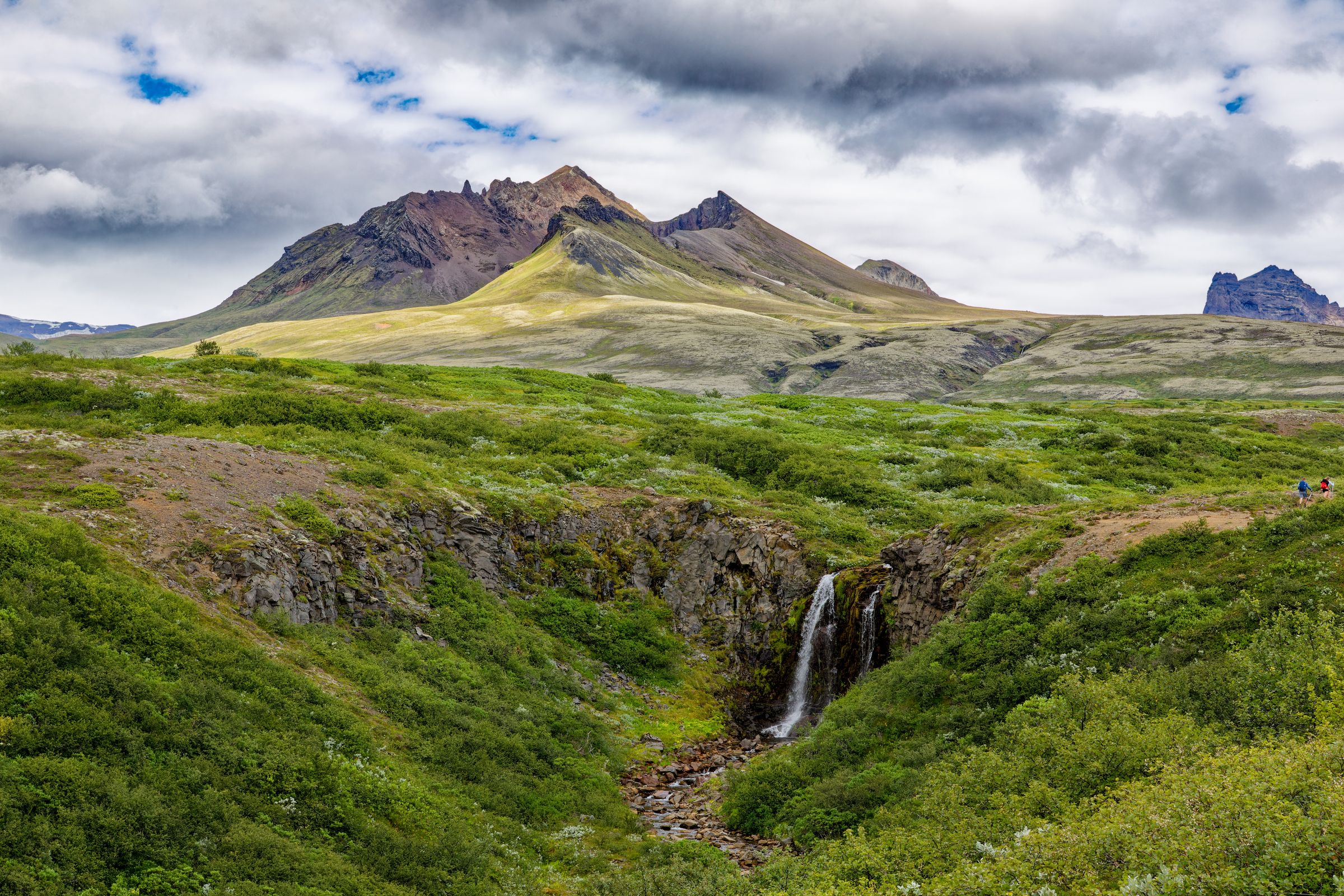 A small waterfall cascades through a lush green landscape with distant mountains under a cloudy sky.