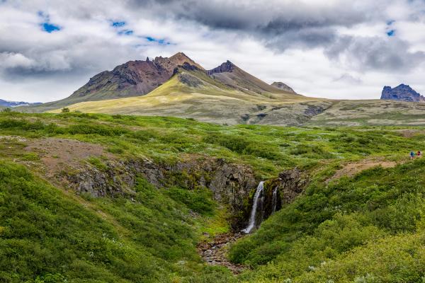 A small waterfall cascades through a lush green landscape with distant mountains under a cloudy sky.