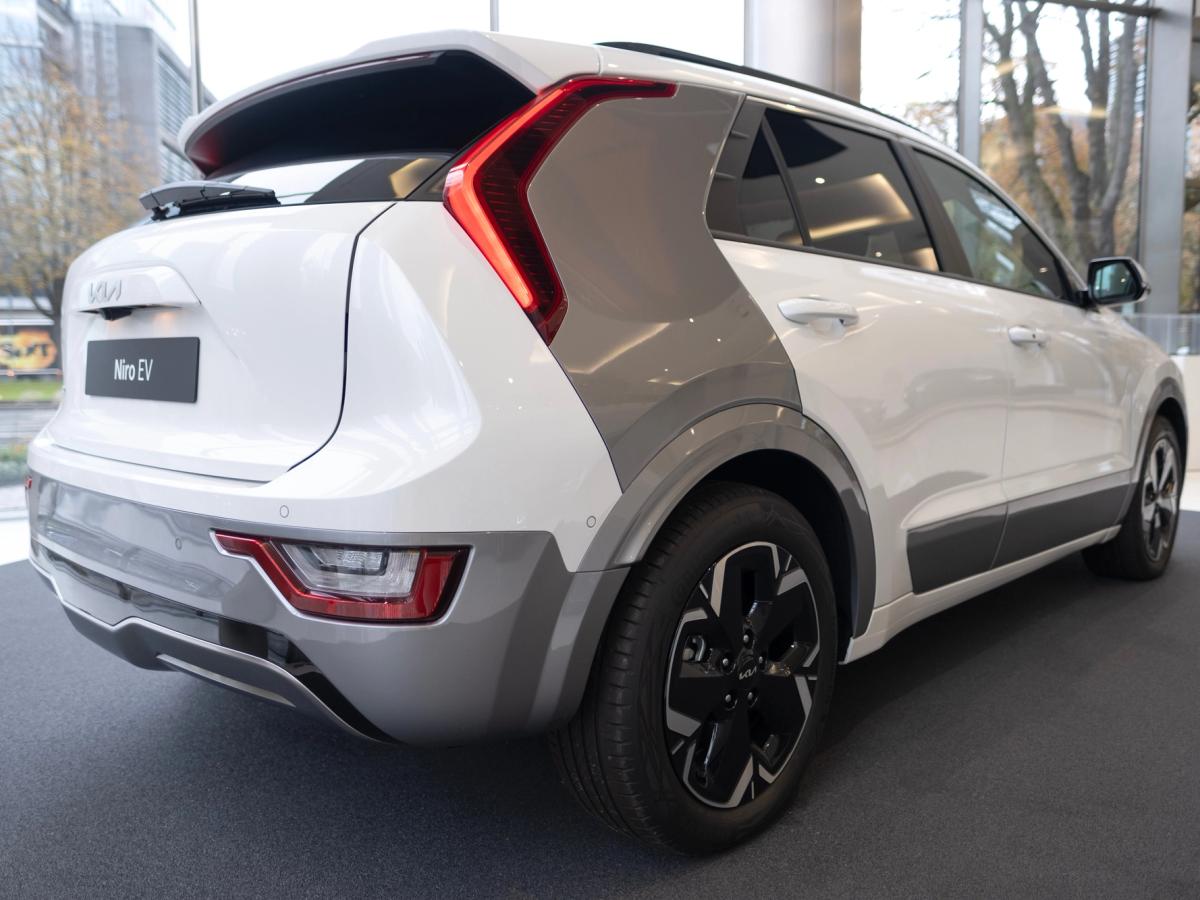 Niro EV séð aftan frá hlið Hvítur Kia Niro EV séður aftan frá hlið í sýningarsal, með gráu þili á C-stoð og afturljósum