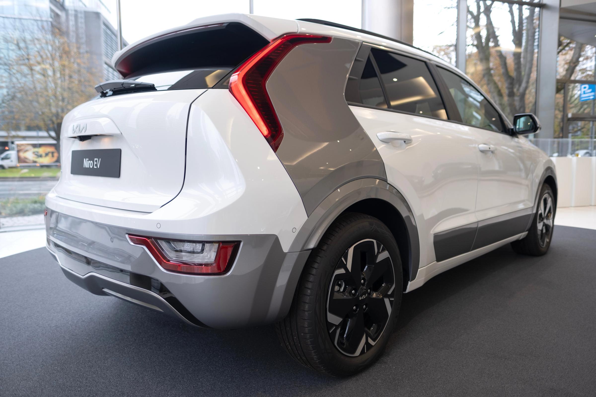 Niro EV séð aftan frá hlið Hvítur Kia Niro EV séður aftan frá hlið í sýningarsal, með gráu þili á C-stoð og afturljósum