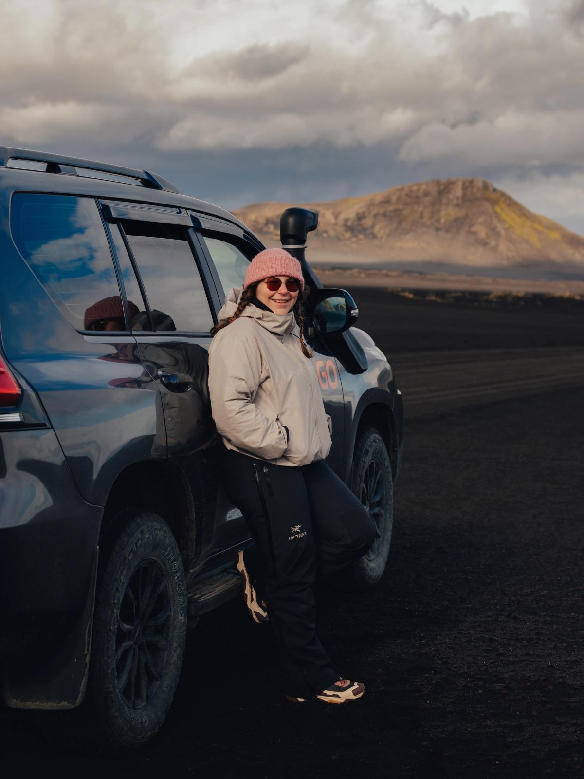 A smiling woman in a pink beanie and sunglasses leans against a dark off-road SUV in a barren volcanic landscape under a cloudy sky.