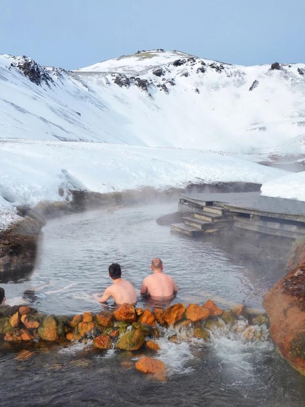 Three people soaking in a steaming hot spring in a snowy mountain valley with a wooden boardwalk.