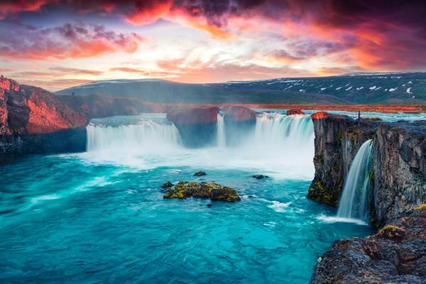 Dramatic sunset sky over a wide waterfall of vivid turquoise water and dark rocky cliffs.