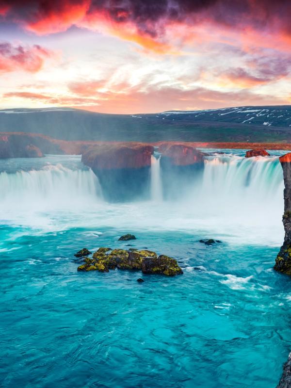 A powerful waterfall plunges into turquoise water amidst rocky cliffs under a vibrant sunset sky.