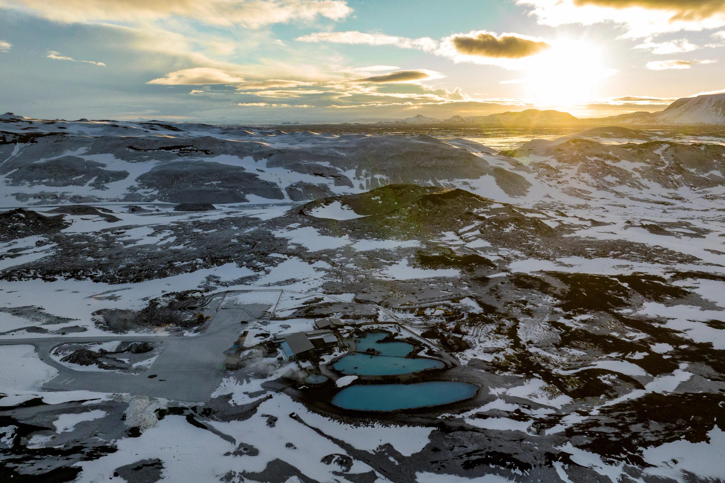 Aerial view of bright blue geothermal pools in a vast, snowy volcanic landscape under a sunset sky.