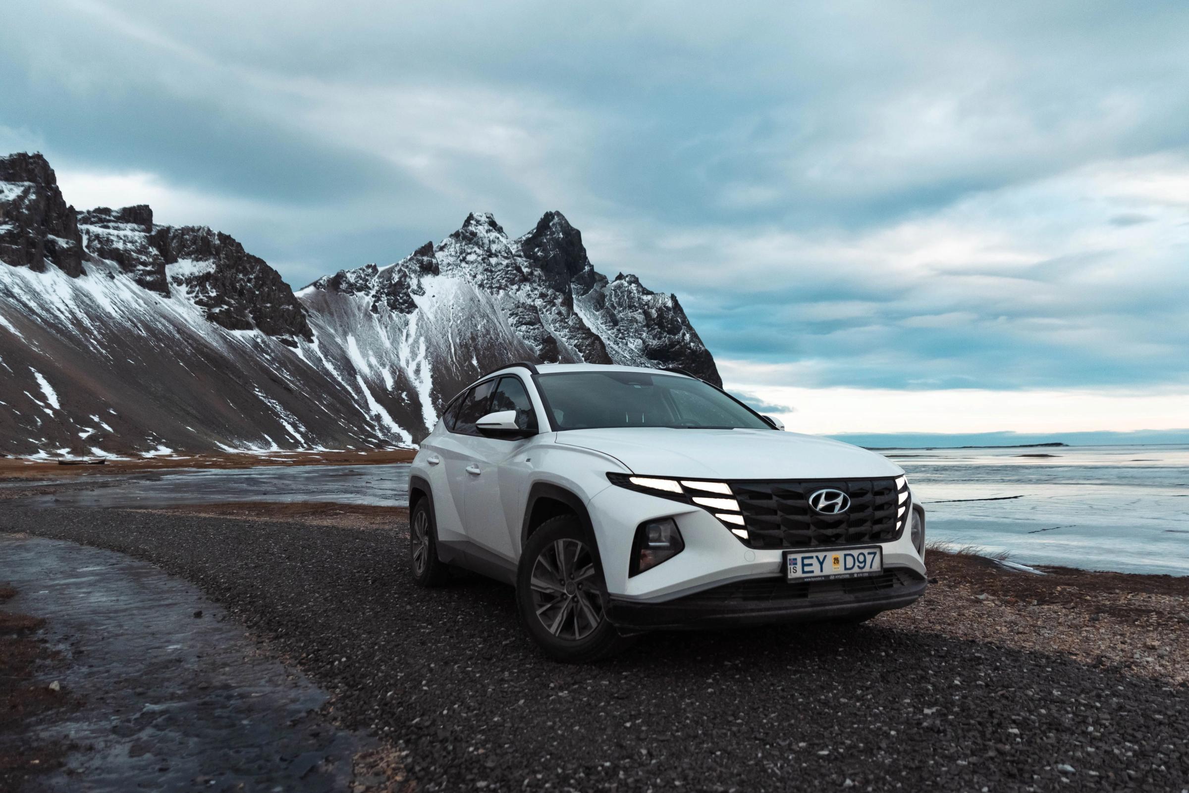 Un SUV Hyundai Tucson blanco en una carretera de grava junto a un agua helada, con montañas nevadas bajo un cielo nublado.