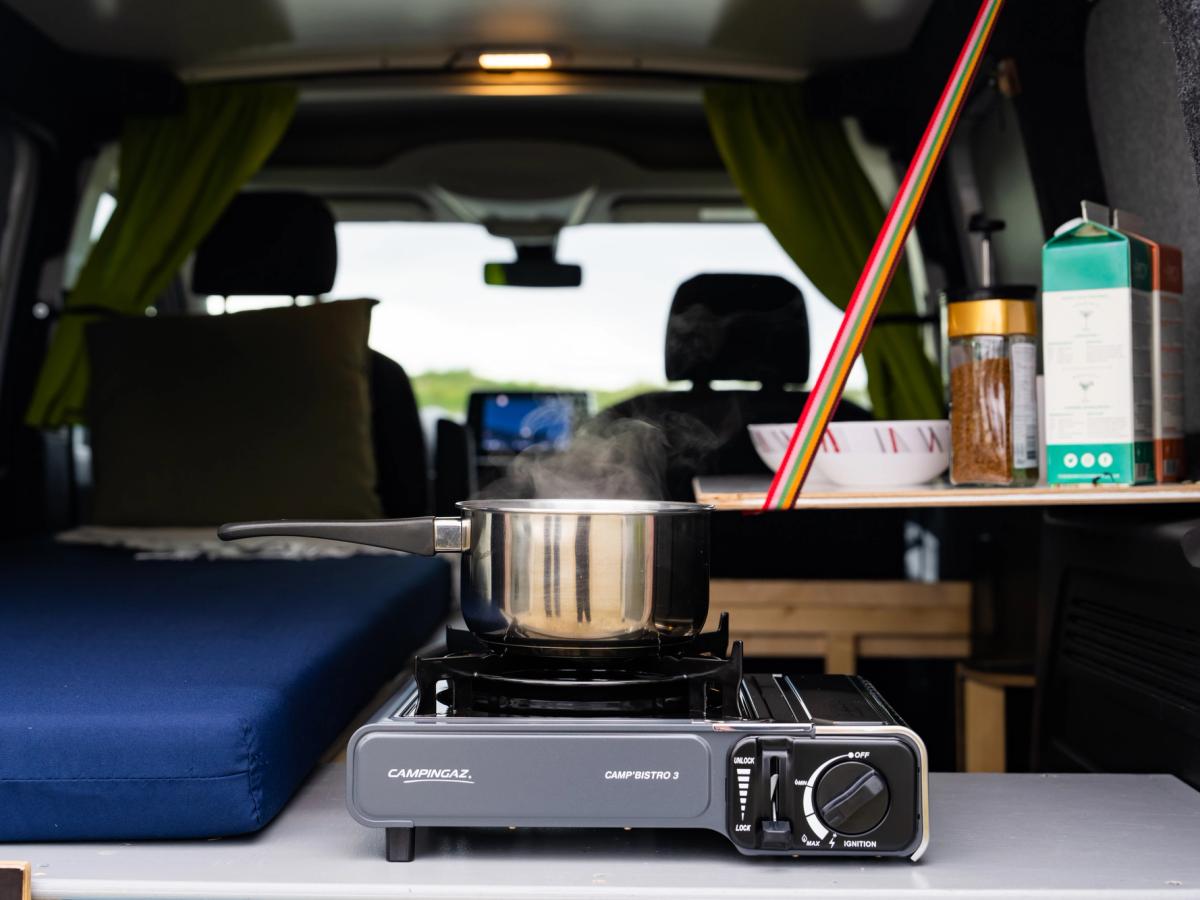Estufa con olla humeante Primer plano de una olla humeante sobre el hornillo de camping, con una mesa, un cuenco y un tarro de café al fondo