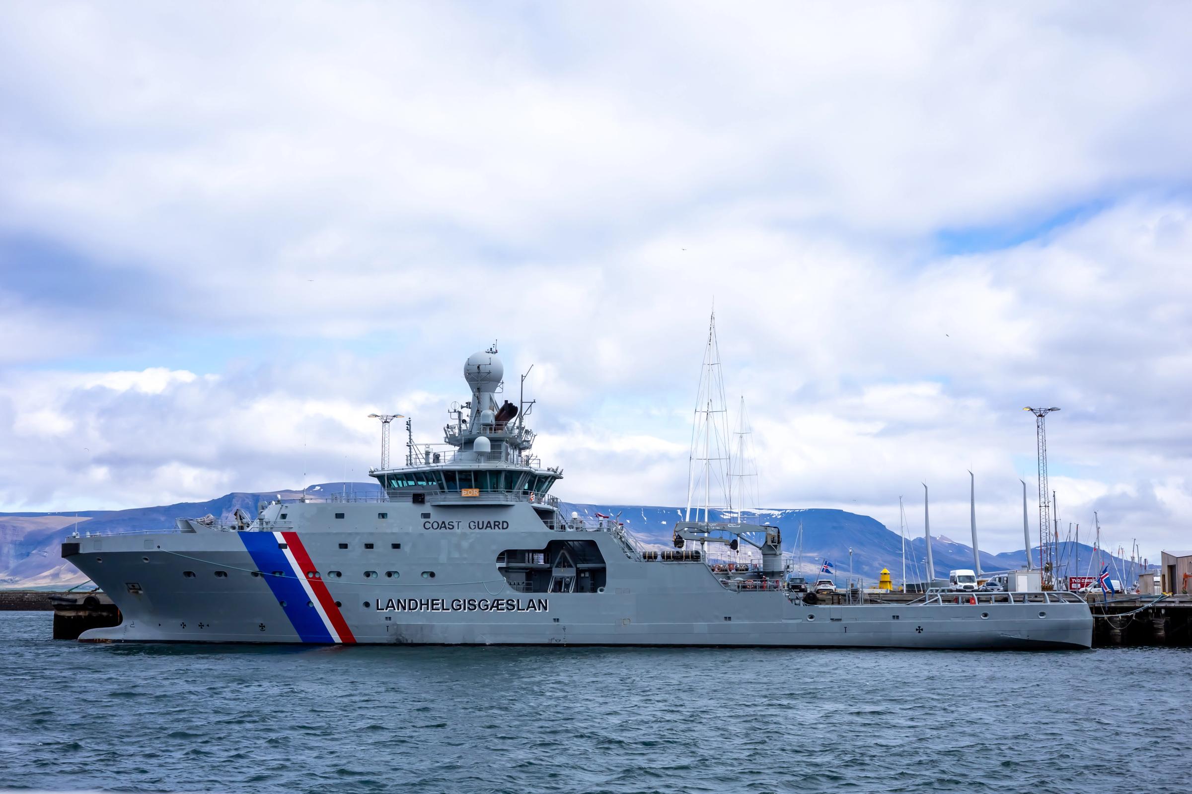 A grey Coast Guard ship with a blue, white, and red stripe on its bow, docked in a harbor.