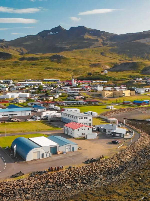 Aerial view of a coastal town with colorful buildings nestled at the base of grassy mountains, next to a rocky shoreline.