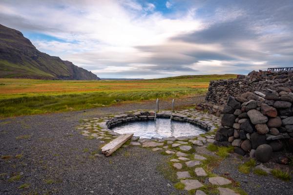 A stone-lined hot spring pool in a green valley with distant mountains and a cloudy sky.