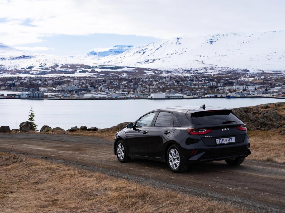Miradouro Costeiro de Ceed Vista traseira de um Kia Ceed estacionado numa estrada de gravilha acima de uma vila costeira, com montanhas ao fundo