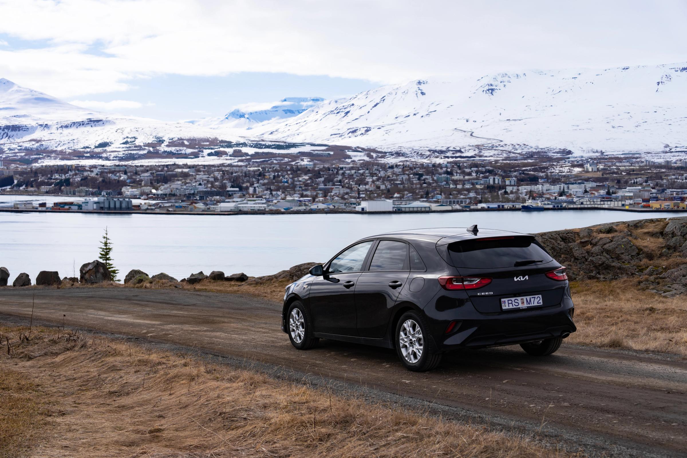 Miradouro Costeiro de Ceed Vista traseira de um Kia Ceed estacionado numa estrada de gravilha acima de uma vila costeira, com montanhas ao fundo
