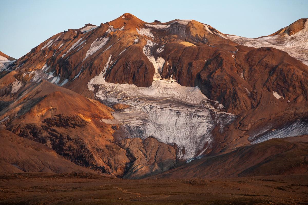 Una montaña de color marrón rojizo con un prominente glaciar blanco y parches de nieve, bañada en una luz cálida.