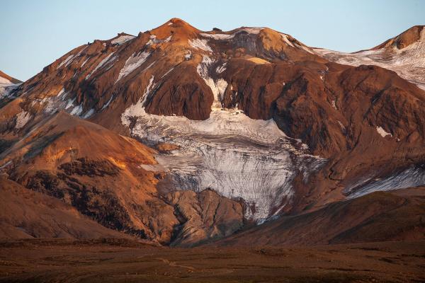 Una montaña de color marrón rojizo con un prominente glaciar blanco y parches de nieve, bañada en una luz cálida.