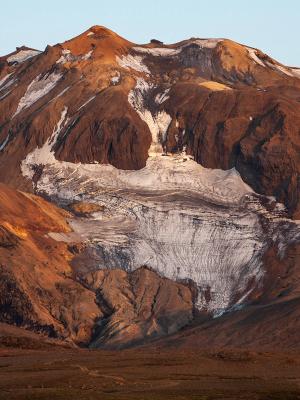 A reddish-brown mountain with a prominent white glacier and patches of snow, bathed in warm light.