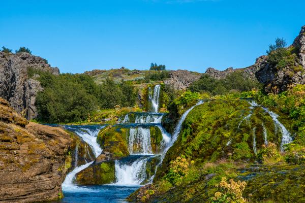 Multi-tiered waterfall flowing over moss-covered rocks into a river under a clear blue sky.