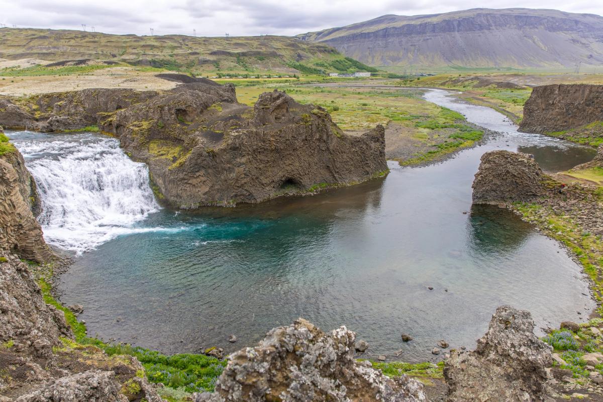 A wide waterfall cascades into a blue-green pool surrounded by dark rock formations and a green valley.