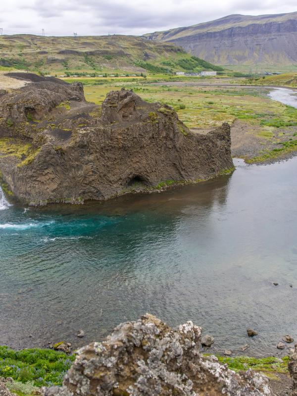 A wide waterfall cascades into a blue-green pool surrounded by dark rock formations and a green valley.