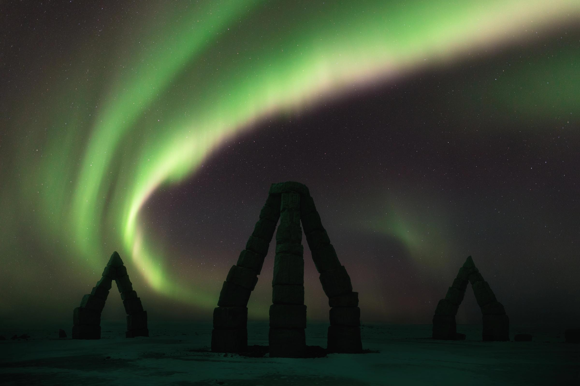 the aurora borealis is shining over a stone arch in the middle of a snowy field .