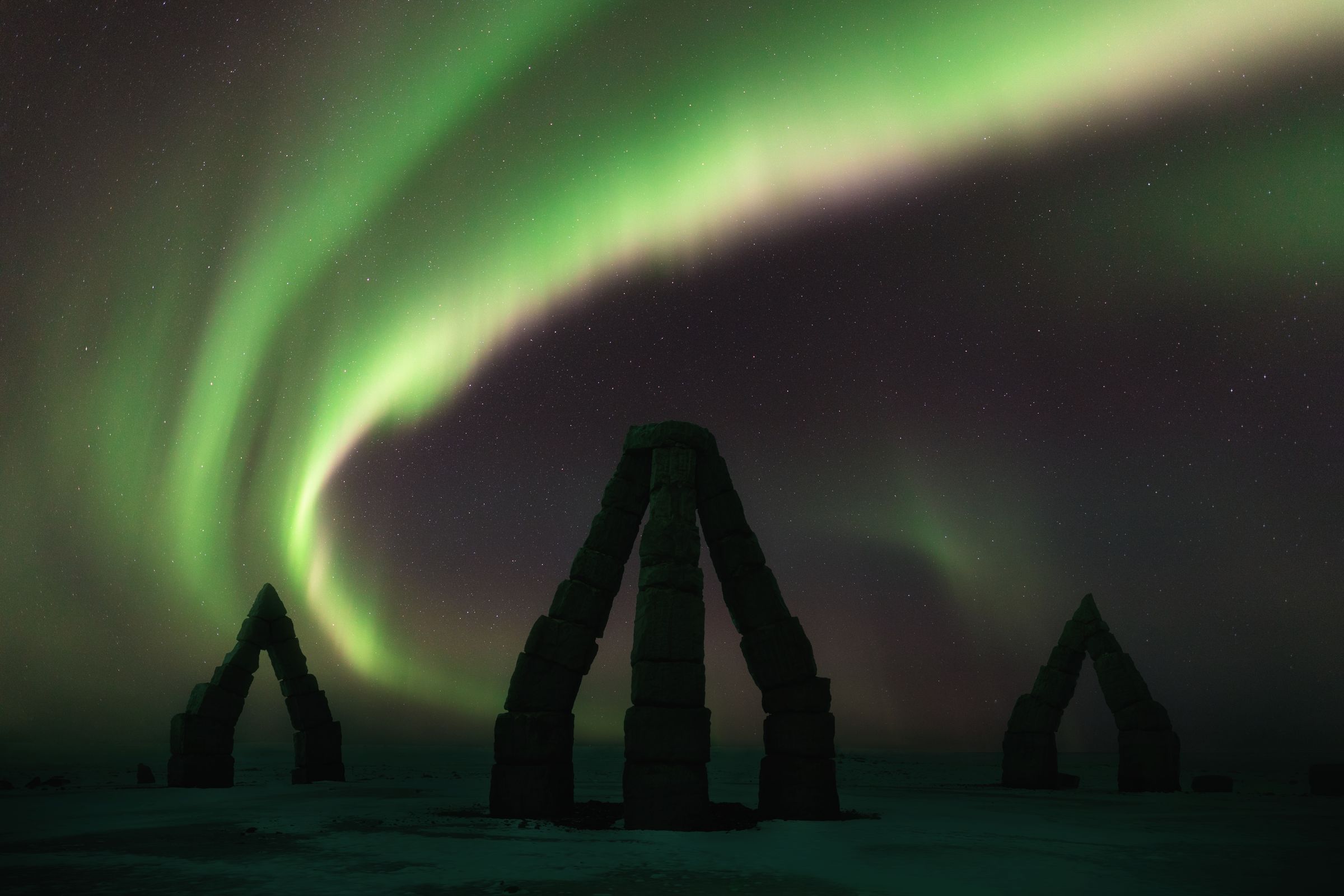 the aurora borealis is shining over a stone arch in the middle of a snowy field .