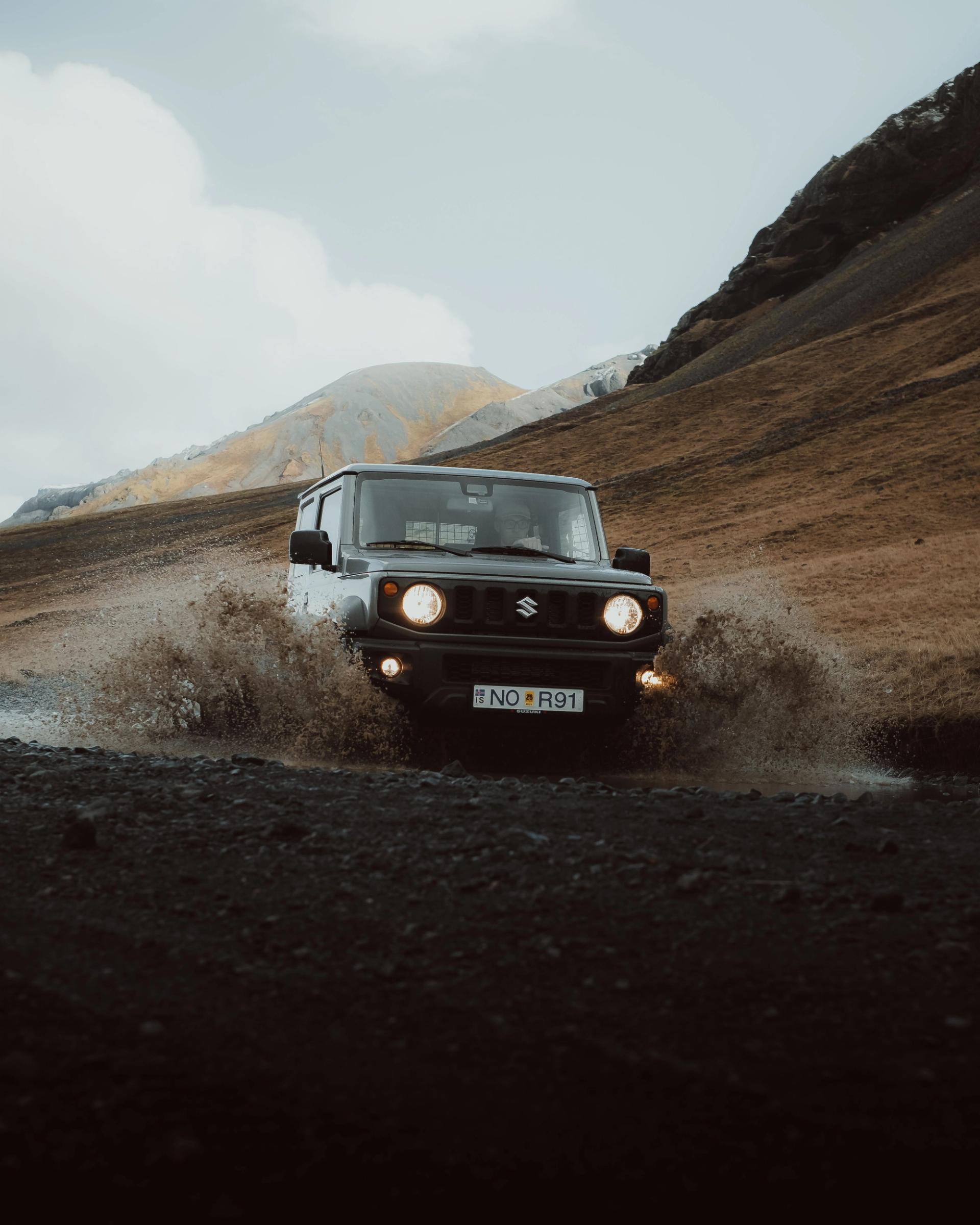 a suzuki jimny is driving through a puddle of water on a dirt road .