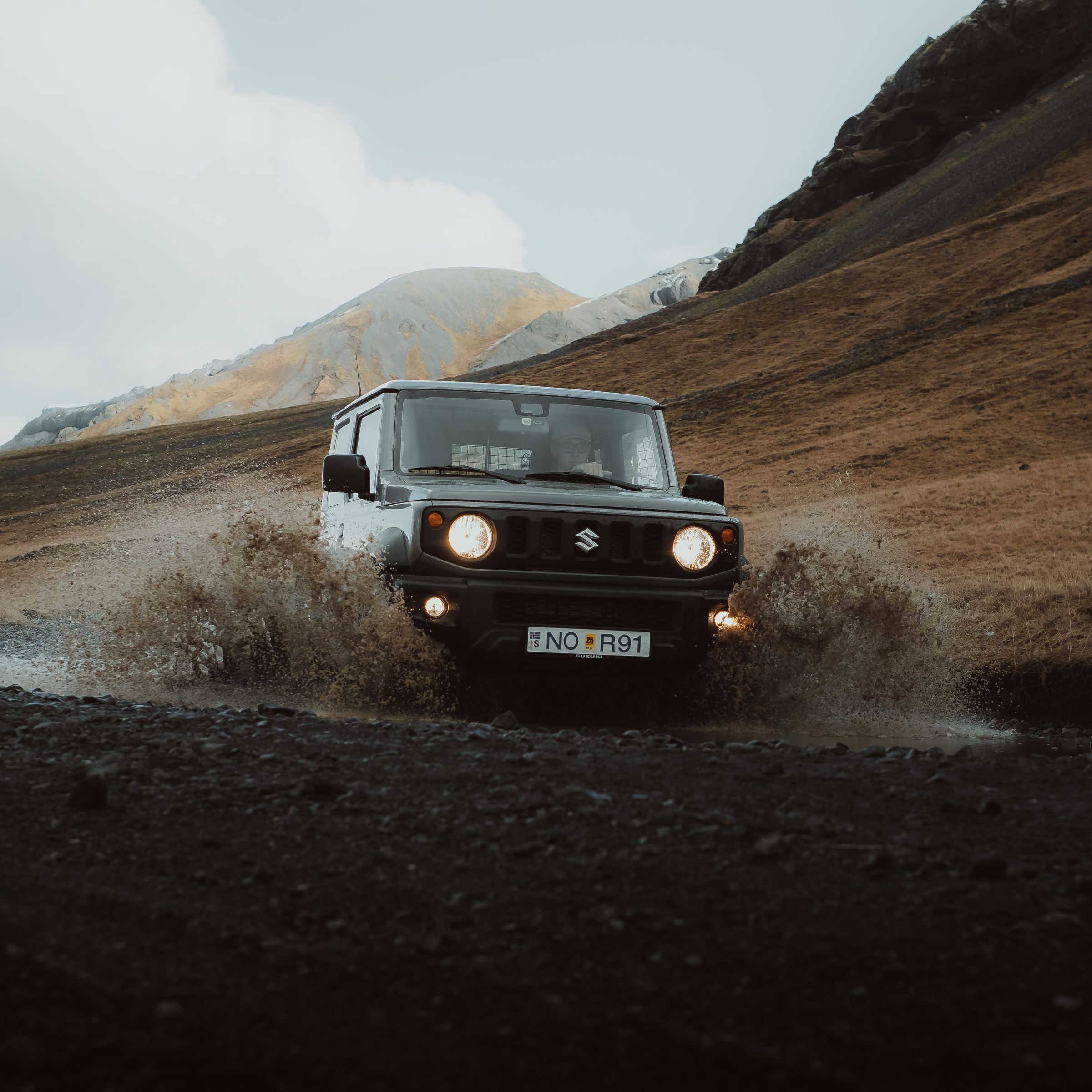 a suzuki jimny is driving through a puddle of water on a dirt road .