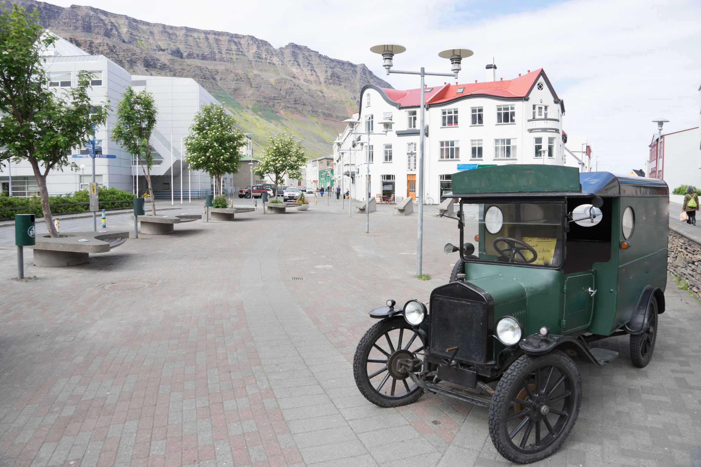A vintage dark green van parked on a brick-paved street with white buildings and a large mountain in the background.