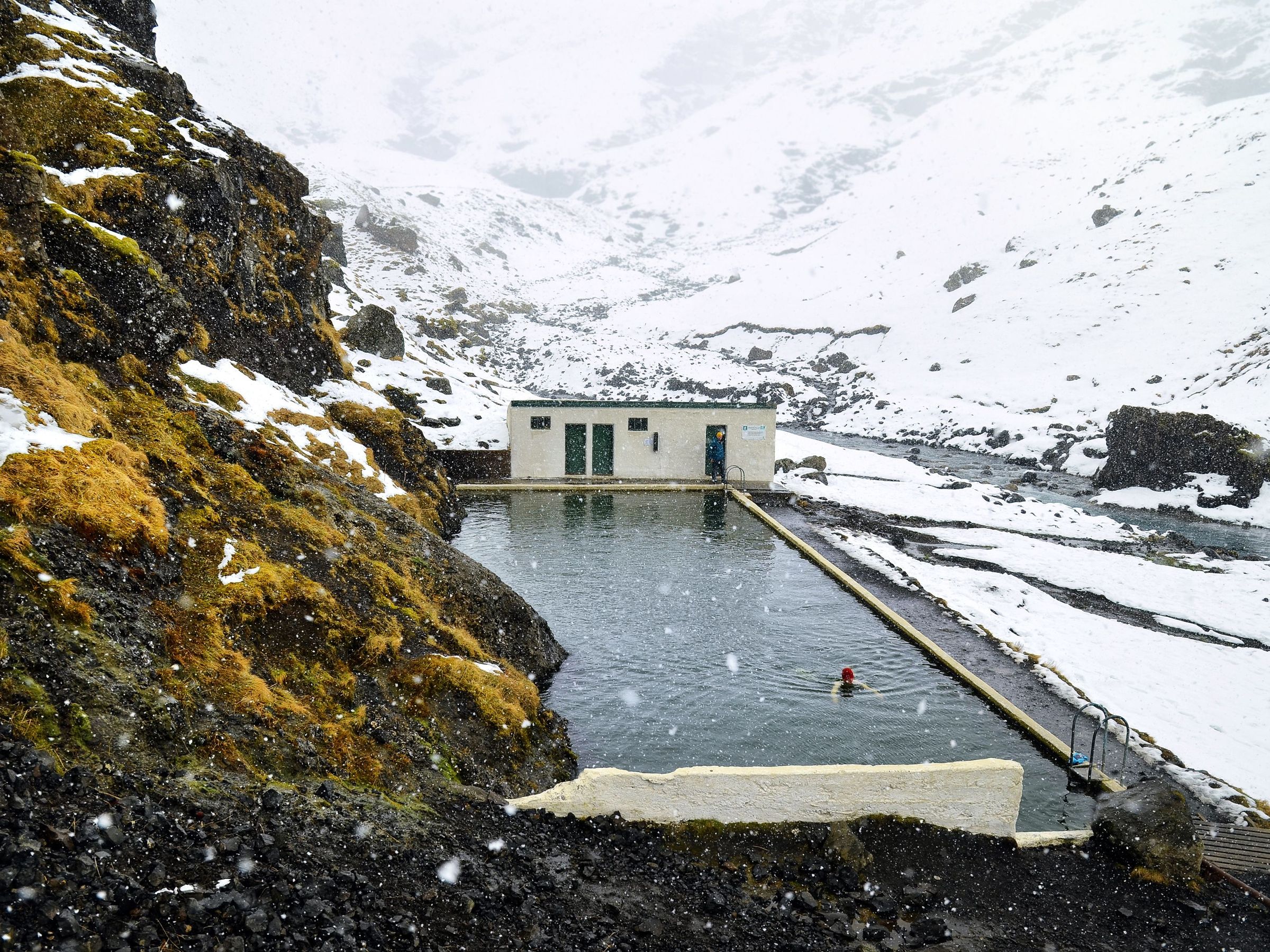 Una persona nada en una piscina termal al aire libre durante una nevada, rodeada de montañas nevadas y un pequeño edificio.