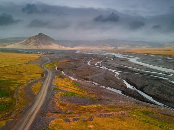A dirt road and braided river flow through yellow-green fields towards a conical mountain and buildings under a dark, cloudy sky.