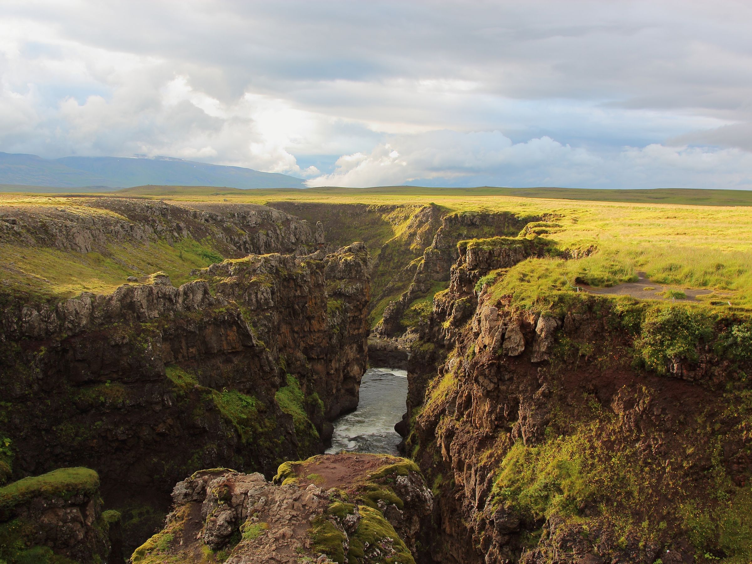 Deep canyon with a river, surrounded by green plains under a cloudy sky.