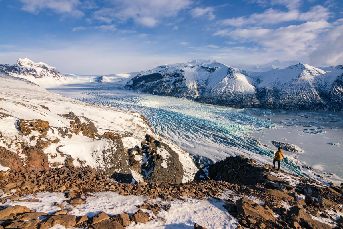una persona está de pie en la cima de una montaña cubierta de nieve mirando un glaciar.